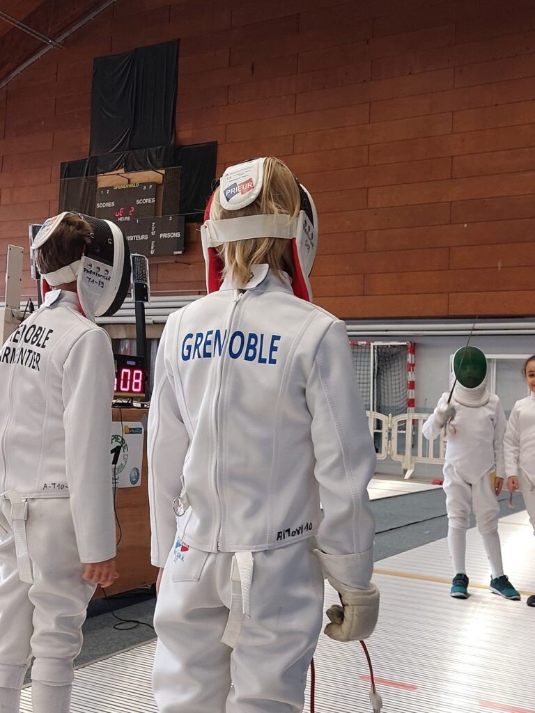 Deux jeunes tireurs vus de dos, veste marquée Grenoble et masque relevé sur la tête, devant le tableau d’affichage des scores.