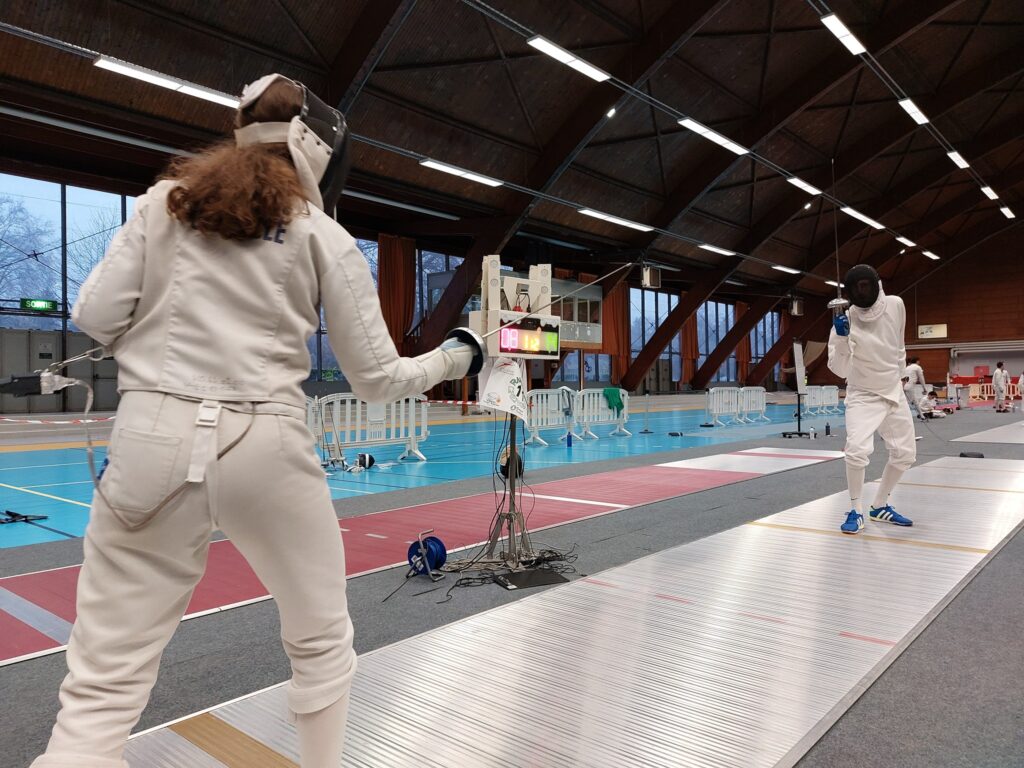 Deux escrimeurs en garde de part et d’autre d’une piste, dans la grande halle au toit de bois ; l’appareil de touche au premier plan.