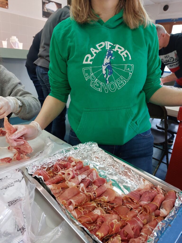 Préparation du repas : un plateau de tranches de jambon roulées, derrière lequel une personne porte le sweat au logo du club.