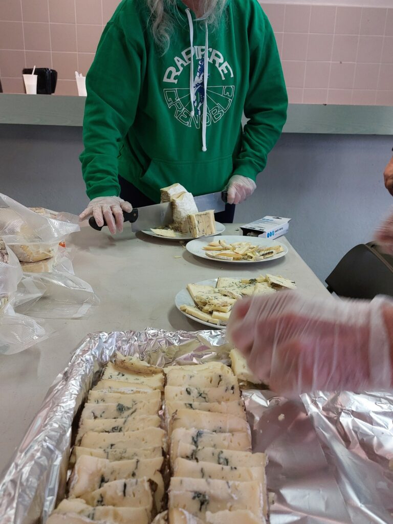 Préparation du repas : une personne en sweat vert du club découpe le bleu du Vercors, les tranches alignées sur un plateau au premier plan.