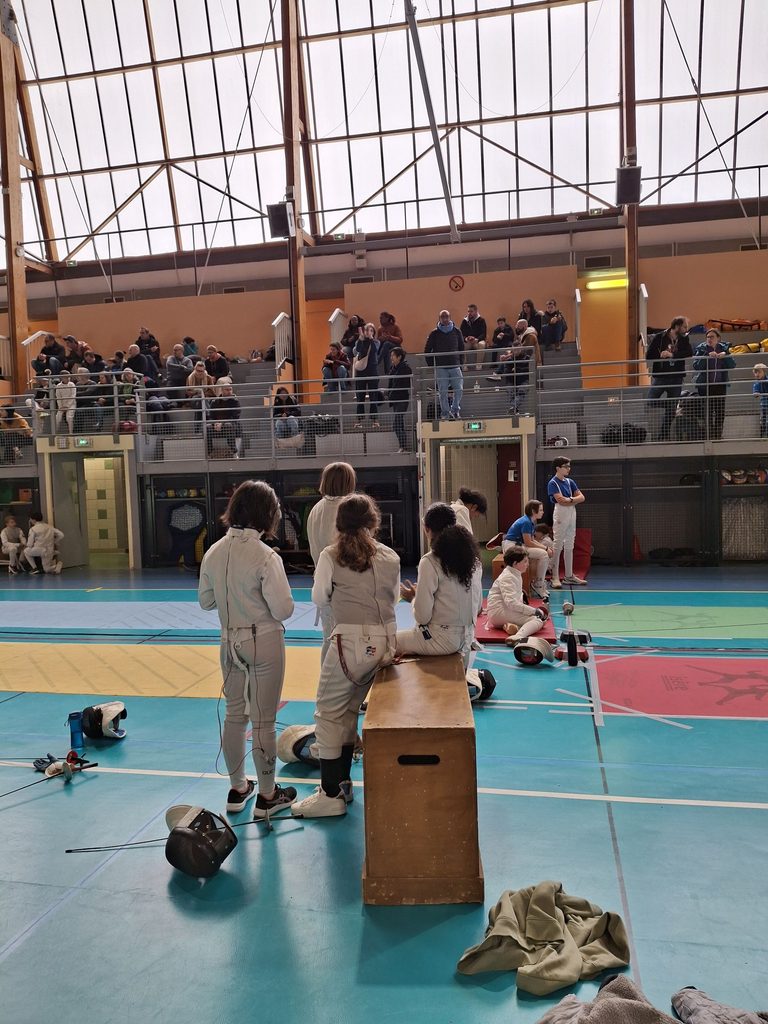 Pendant le tournoi Bayard 2026, de jeunes escrimeurs attendent leur tour au bord de la piste, devant des gradins garnis de parents.