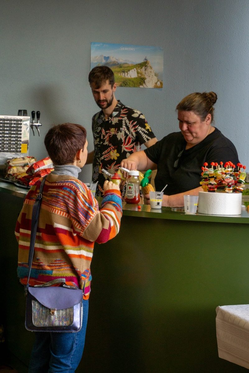 Une bénévole sert une visiteuse au comptoir de la buvette, sous une affiche du Vercors